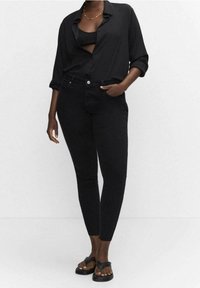 Femme portant un haut noir court, une chemise noire déboutonnée, un jean noir skinny taille haute et des sandales noires, debout contre un mur blanc.