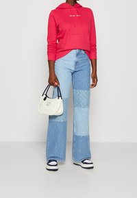Donna che indossa una felpa rossa con cappuccio, jeans a palazzo in patchwork, sneakers bianche e blu navy, mentre tiene una piccola borsa bianca con manici blu navy.