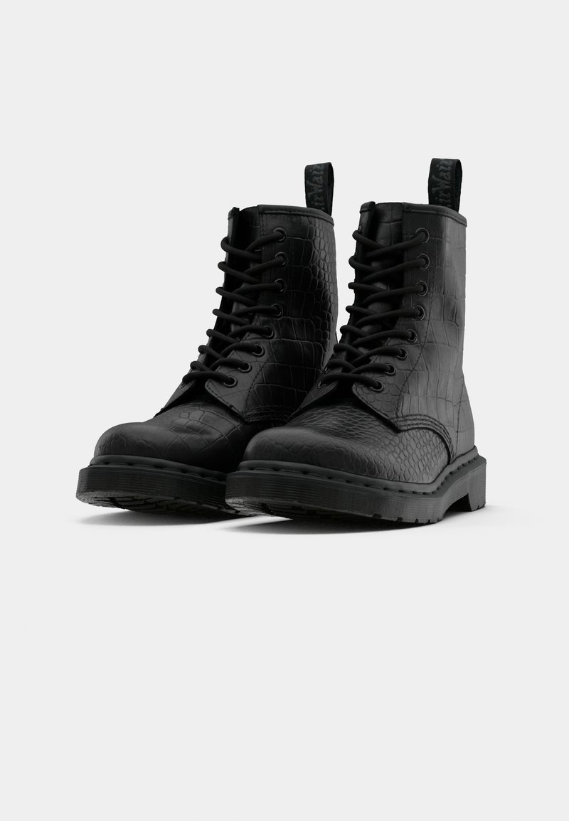 Botas de combate de cuero negro con textura de cocodrilo, suelas gruesas, cordones y tiradores, sobre un fondo blanco liso.