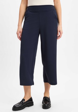 Frau trägt marineblaue, gekürzte, weit geschnittene Hosen und schwarze Loafer, steht vor einem schlichten weißen Hintergrund.