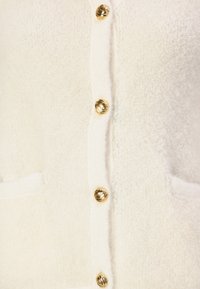 Gilet en polaire blanche avec un devant à boutons, orné de boutons ronds dorés et de deux poches avant. Texture douce et apparence chaleureuse.
