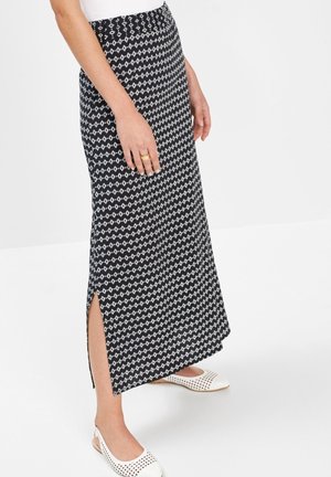 Donna che indossa una gonna lunga nera e bianca con motivo decorativo e spacco laterale, e ballerine slingback bianche traforate, in piedi su un pavimento bianco.