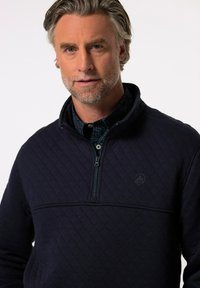 Sudadera acolchada en azul marino con cuello con cremallera, que presenta un panel texturizado en contraste y un detalle del logo en el pecho. Acabado de tela suave.