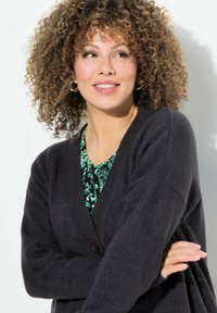 Schwarze Strickjacke mit tiefem V-Ausschnitt und gerippten Bündchen, getragen über einer grün gemusterten Bluse. Das Model hat lockige Haare und große Ohrringe.