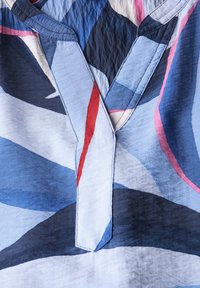 Blusa multicolore con motivi geometrici in tonalità di blu, rosso e rosa. Design con scollo a V, leggermente testurizzato e con cuciture visibili.