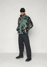 Fleece jakna v zeleni barvi s roza listnatim vzorcem, ki vključuje žep na prsih, zadrgo in rebraste manšete; v kombinaciji s črnimi hlačami in čevlji.