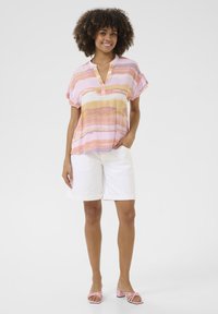Blusa a maniche corte di un delicato rosa con righe orizzontali gialle e viola, abbinata a pantaloni corti bianchi e sandali con tacco rosa.