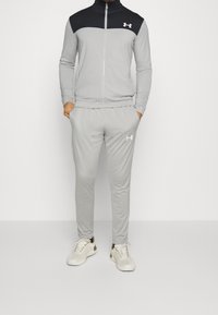 Homme portant un survêtement Under Armour gris clair avec un panneau noir sur la poitrine et des baskets blanches, debout les mains dans les poches sur fond blanc.
