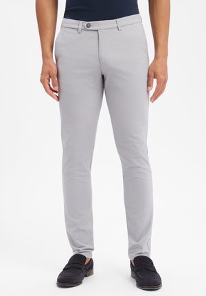 Mann trägt hellgraue Slim-Fit-Hosen mit Knopf und Reißverschluss, kombiniert mit schwarzen Loafers und einem dunklen Oberteil, steht vor weißem Hintergrund.