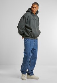 Sudadera con capucha gris oscuro, con bolsillo frontal y logo negro. Combinada con jeans de pierna ancha azul claro y zapatillas de color claro.
