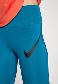Leggings de sport turquoise en matériau élastique, présentant un logo Nike noir sur la cuisse et une texture lisse avec des coutures plates.