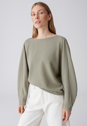 Vrouw met een lichte huid en lang blond haar, gekleed in een saliegroene geribbelde trui en witte hoge taille broek, staand tegen een grijze achtergrond.