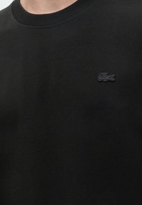 Gros plan d'une personne portant un t-shirt noir à col rond avec un petit logo crocodile noir brodé sur la poitrine gauche.