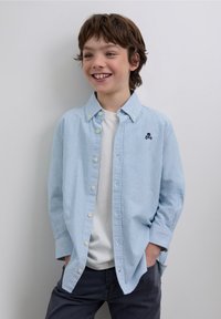 Vaaleansininen, raidallinen nappipaita kauluksella, jossa on pieni kirjottu kallo ja ristiluut rinnassa, yhdistettynä valkoiseen t-paitaan.