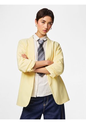 Young person with short dark hair, wearing a yellow blazer, white shirt, striped tie, and dark jeans, standing with arms crossed against white background.