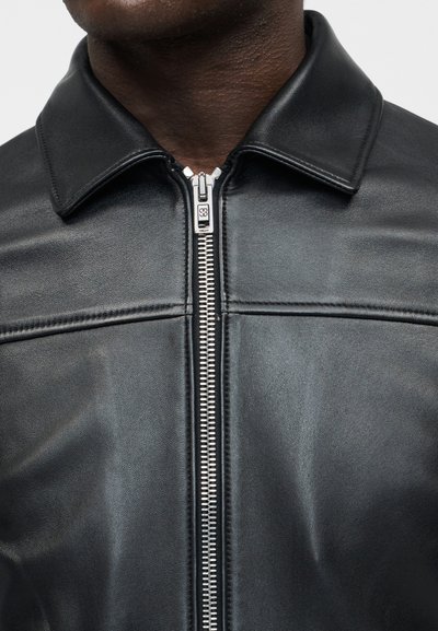 Κοντινό πλάνο ενός μαύρου δερμάτινου μπουφάν με ασημένιο φερμουάρ μπροστά και διπλωμένο γιακά, που φοριέται από ένα άτομο με σκουρόχρωμο δέρμα.