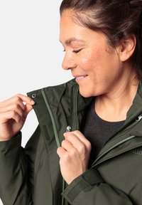 Groene waterdichte jas met een ritssluiting aan de voorkant, handzakken en klittenbandaccenten. Glad oppervlak en sportief ontwerp, gedragen over een donker shirt.