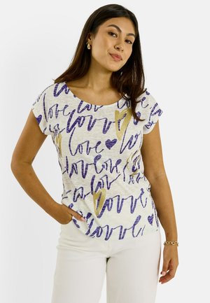 Mujer vistiendo una camiseta blanca con texto "love" en púrpura y dorado y diseños de corazones, combinada con pantalones blancos, de pie con una mano en el bolsillo.