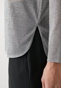 Zilveren, metallic blouse met lange mouwen en een subtiele glans, gebogen zoom en een zijsplit, gecombineerd met een zwart kledingstuk eronder.
