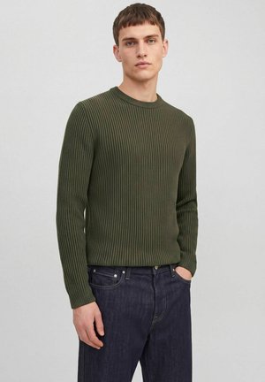 Jeune homme aux cheveux bruns courts portant un pull côtelé vert olive et un jean bleu foncé, debout avec une main dans la poche.