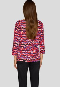 Multicolored top featuring a zigzag pattern in reds, purples, and whites; long sleeves; and a relaxed fit, paired with dark pants.