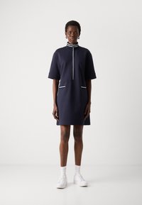 Navy short-sleeve dress with a mock neck, zip closure, and white stripe accents on pockets and collar. Smooth textured fabric. White shoes.