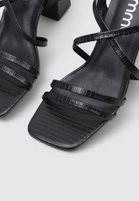 Sandalias de tacón negro con un diseño de punta cuadrada. La textura tiene un estampado con un relieve parecido al de un cocodrilo. Presenta múltiples tiras delgadas.