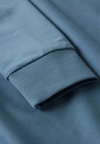 Blå sweatshirtärm med ribbade manschetter, som visar tygstrukturen och sömdetaljer i närbild.