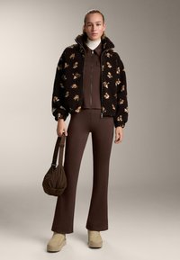 Femme portant une veste marron zippée et un pantalon avec des chaussures compensées beige, tenant un sac marron, debout devant un fond uni.
