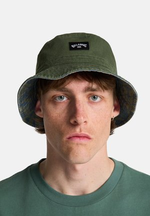 Młody mężczyzna o jasnej karnacji i niebieskich oczach, noszący oliwkowozielony kapelusz typu bucket marki Billabong oraz dopasowaną zieloną koszulkę z okrągłym dekoltem.