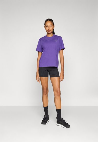 Lila rövid ujjú pamut póló fekete sportos rövidnadrággal. A modell fekete cipőt és zoknit visel, semleges háttér előtt áll.