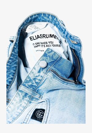 Lyseblå denimjakke med falmet tekstur; har glidelås, trykknapp og en logopatch på lommen. Innvendig etikett synlig.