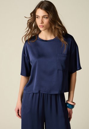 Ung kvinde med bølget brunt hår iført en løs mørkeblå top med korte ærmer og matchende bukser med vide ben, accessorized med chunky armbånd.