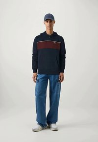 Navy hoodie with red stripe and white accent, paired with wide blue denim cargo pants. Model wears a gray cap and white sneakers.