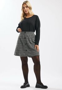 Schwarzes Langarmoberteil kombiniert mit einem karierten Rock mit Knopfverschluss, strukturiertem Stoff und schwarzen blickdichten Strumpfhosen. Schwarze Schuhe runden das Outfit ab.