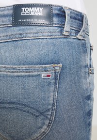 Modré džíny z denimu s koženou nášivkou označenou "TOMMY JEANS" s bílým textem. Malá trojbarevná logo ozdoba na zadní kapse.