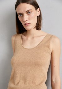 Débardeur en tricot beige sans manches avec un encolure dégagée. Texture douce, design ajusté et détails minimalistes. Convient pour être porté en superposition ou seul.