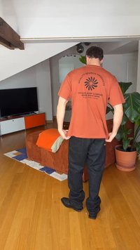 Άνδρας στέκεται στο σαλόνι, φοράει πορτοκαλί μπλούζα Ripcurl Surf Company, μαύρο παντελόνι, κοντά σε καναπέ με πορτοκαλί κουβέρτα και μεγάλο γλαστρικό φυτό.