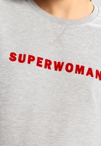 Šedá mikina s odvážným červeným vyšitým nápisem "SUPERŽENA". Bavlněný materiál s kulatým výstřihem a texturovanou tkaninou.