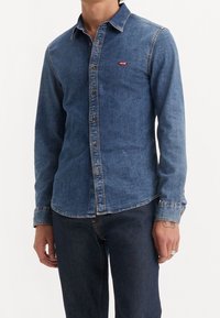 Camicia di jeans blu scuro con colletto a punta, maniche lunghe, chiusura frontale con bottoni e un piccolo logo rosso sul lato sinistro del petto.