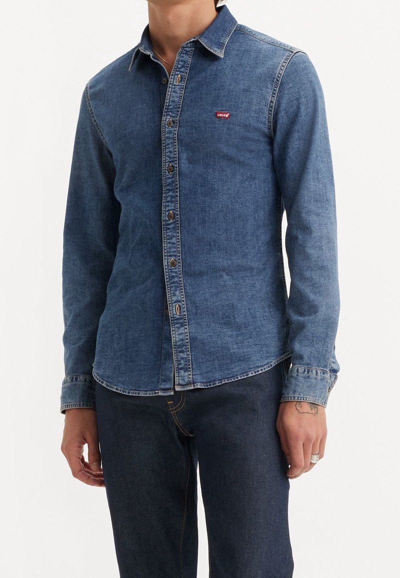 Camicia di jeans blu scuro con colletto a punta, maniche lunghe, chiusura frontale con bottoni e un piccolo logo rosso sul lato sinistro del petto.