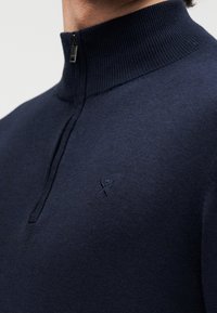 Pull en maille bleu marine avec col zippé, texture côtelée et petit logo brodé sur la poitrine.