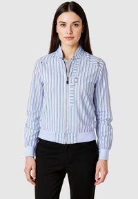 Mujer vistiendo una chaqueta con cremallera de rayas verticales en azul claro y blanco, con puños y cuello acanalados, combinada con pantalones negros, de pie frente a un fondo gris.