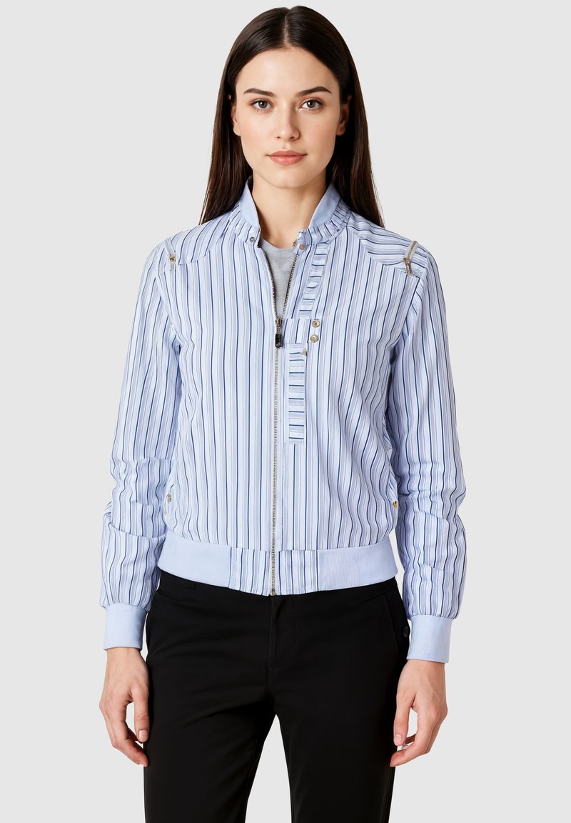 Mujer vistiendo una chaqueta con cremallera de rayas verticales en azul claro y blanco, con puños y cuello acanalados, combinada con pantalones negros, de pie frente a un fondo gris.