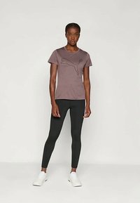 Koszulka z krótkim rękawem w kolorze mauve z ciemnym logo Puma, w parze z czarnymi legginsami i białymi trampkami, na neutralnym tle.