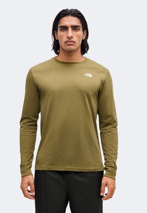 Homme portant une chemise à manches longues vert olive avec le logo The North Face sur la poitrine, debout devant un fond clair uni.