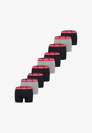 Hemdloser Mann mit verschränkten Händen, der schwarze Levi's Boxershorts mit rotem Bund vor einem einfarbigen weißen Hintergrund trägt.