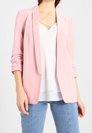 Donna che indossa un blazer rosa chiaro con maniche arricciate, sopra una maglietta bianca con spalline e jeans blu, in piedi di fronte a uno sfondo neutro.