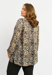 Langärmlige, lockere Bluse in Beige mit schwarzem Blumenmuster, mit abgerundetem Saum und seitlichen Schlitzen für Bewegungsfreiheit.