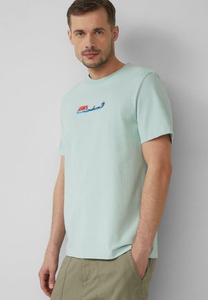Light mint green t-shirt made of cotton, featuring a small red and blue "JAWS" graphic and wave design on the front, short sleeves.